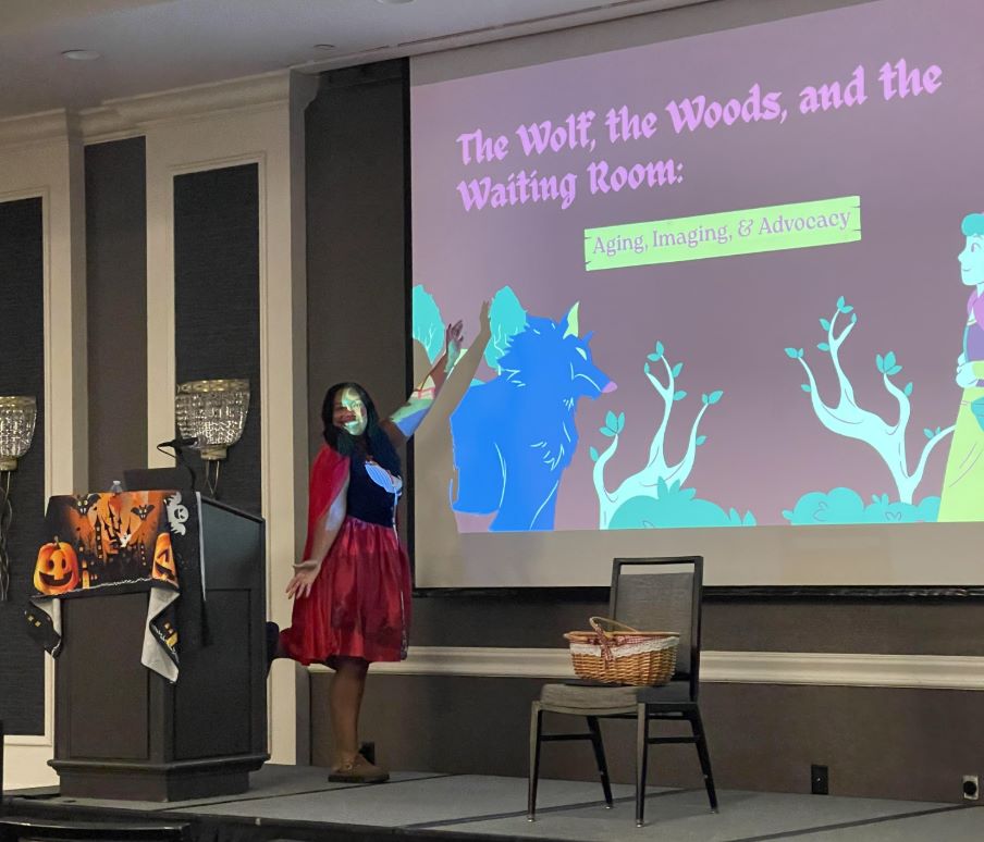 clinical Coordinator Charlie Downing in a red riding hood costume standing on stage presenting The Wolf, the Woods and the Waiting Room: Aging, Imaging and Advocacy.