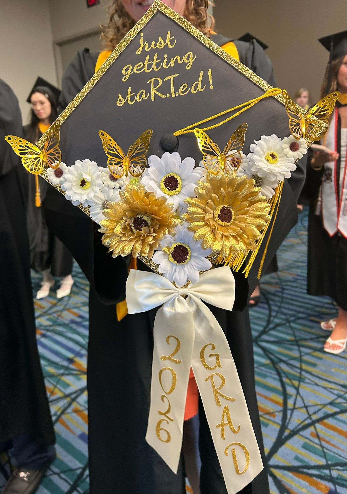 Graduation cap decorated in gold butterflies and white and yellow flowers that reads just getting staRTed 2026 grad